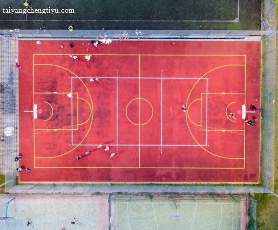 An aerial view of a sports court with a vibrant red surface and yellow lines marking different sections for basketball and soccer. Several people are present on the court, some engaged in activities, while others are standing along the perimeter. The court is enclosed by a metal fence and surrounded by grass on all sides.